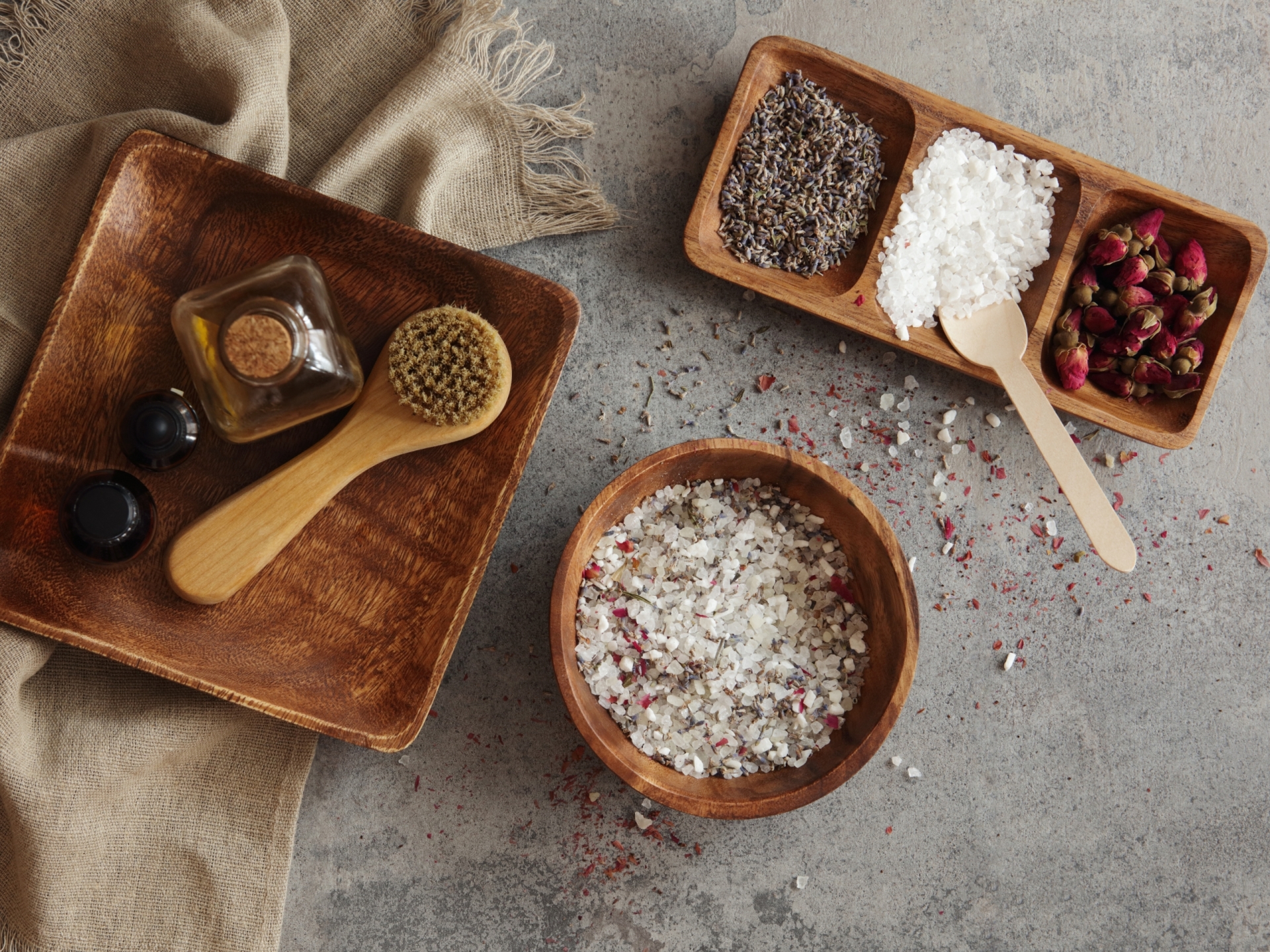 Overhead view of spa ingredients: lavender, sea salt, dried roses, body brush, and essential oils on wooden trays.