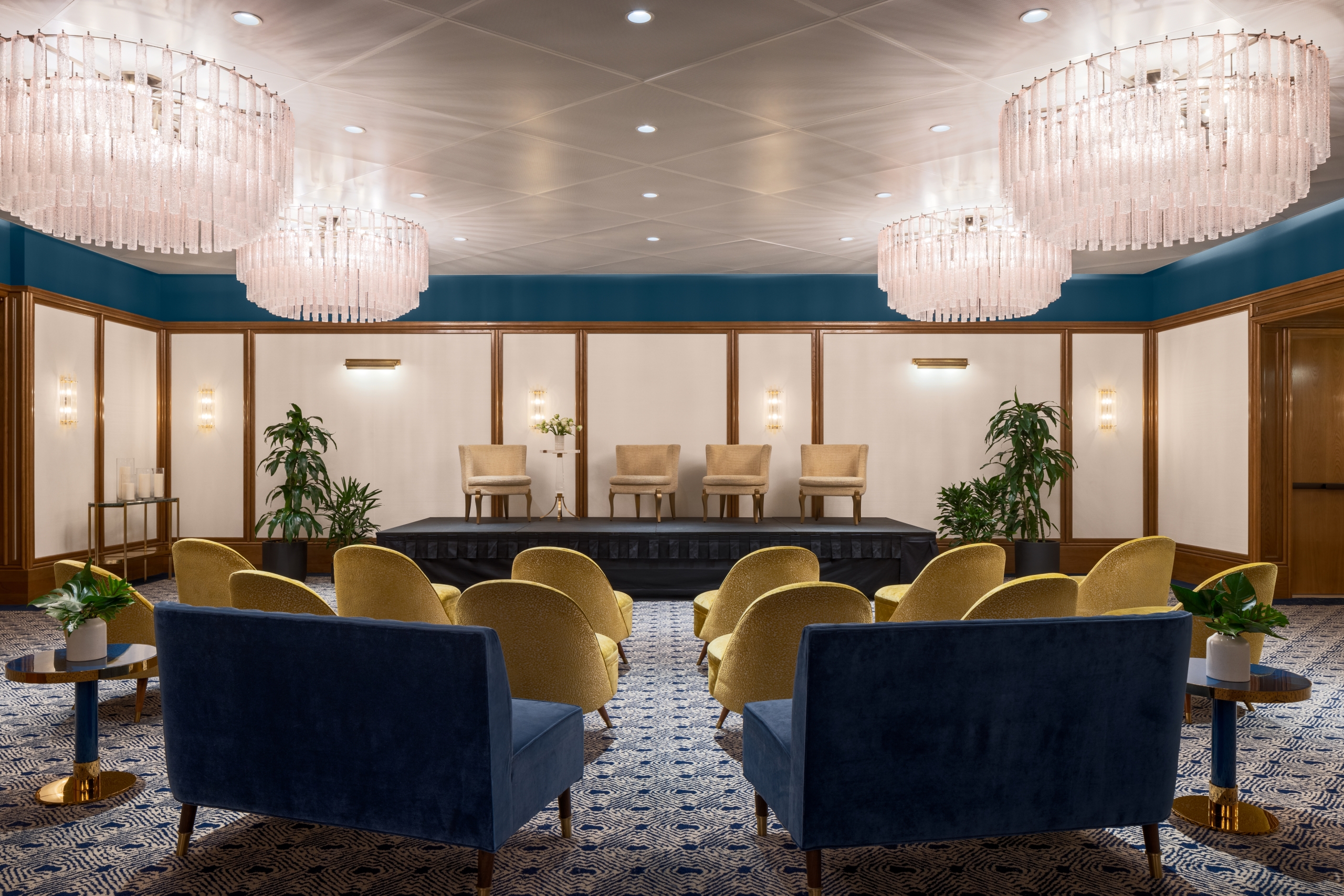 A meeting space in The Sun Rose Ballroom, featuring a patterened blue and tan carpet and four large chandeliers, with plush velvet chairs and couches set up in front of a stage with chairs upon it.