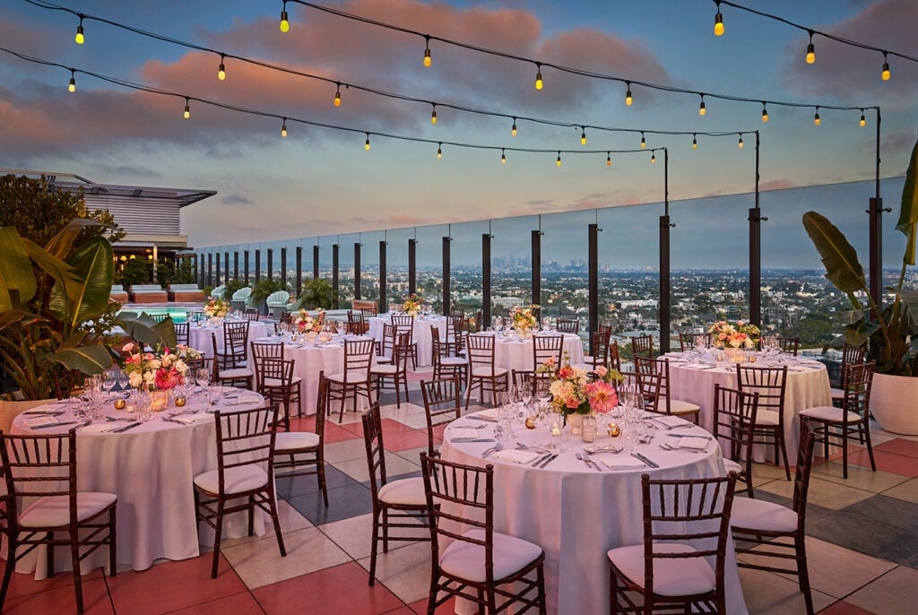 The Rooftop Pool area with tables and chairs set up for a wedding reception at The Sun Rose, with overhead string lighting and sunset views of the los angeles skyline