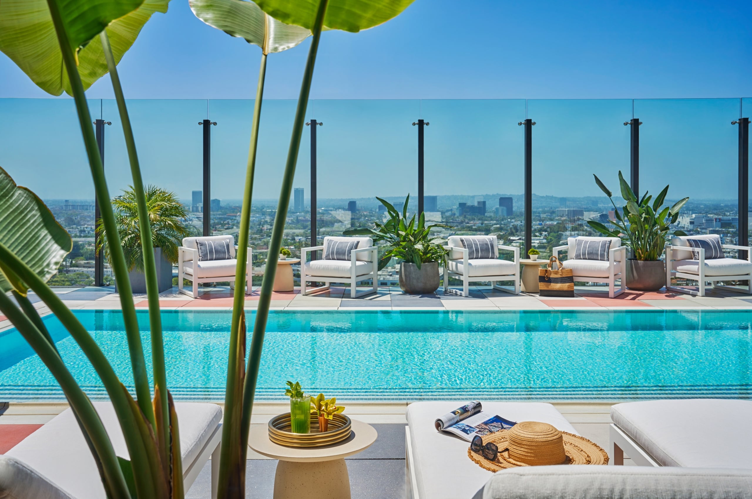 A view of the rooftop pool deck with clear water and sun loungers. The Los Angeles skyline is visible beyond the glass barrier.