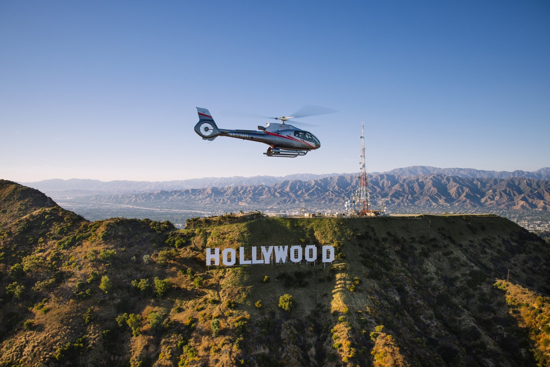 An aerial view of a helicopter flying over the HOLLYWOOD sign on a dry hill with mountains in the background at sunset.