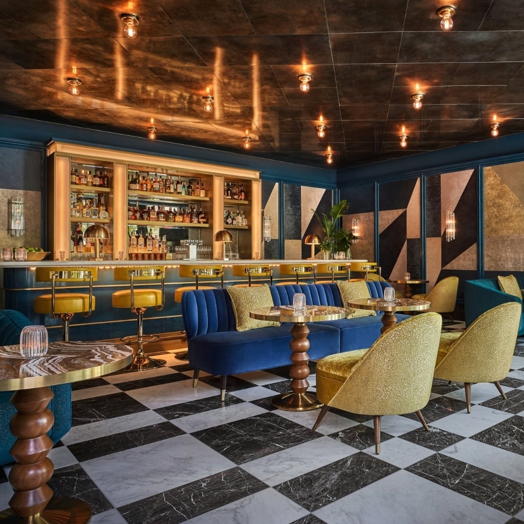 Ritual, the Sun Rose Lobby bar showcasing comfortable bar stools and intimate seating areas with plush couches and lounge chairs upon a checkered marbled floor, bathed in warm ambient lighting