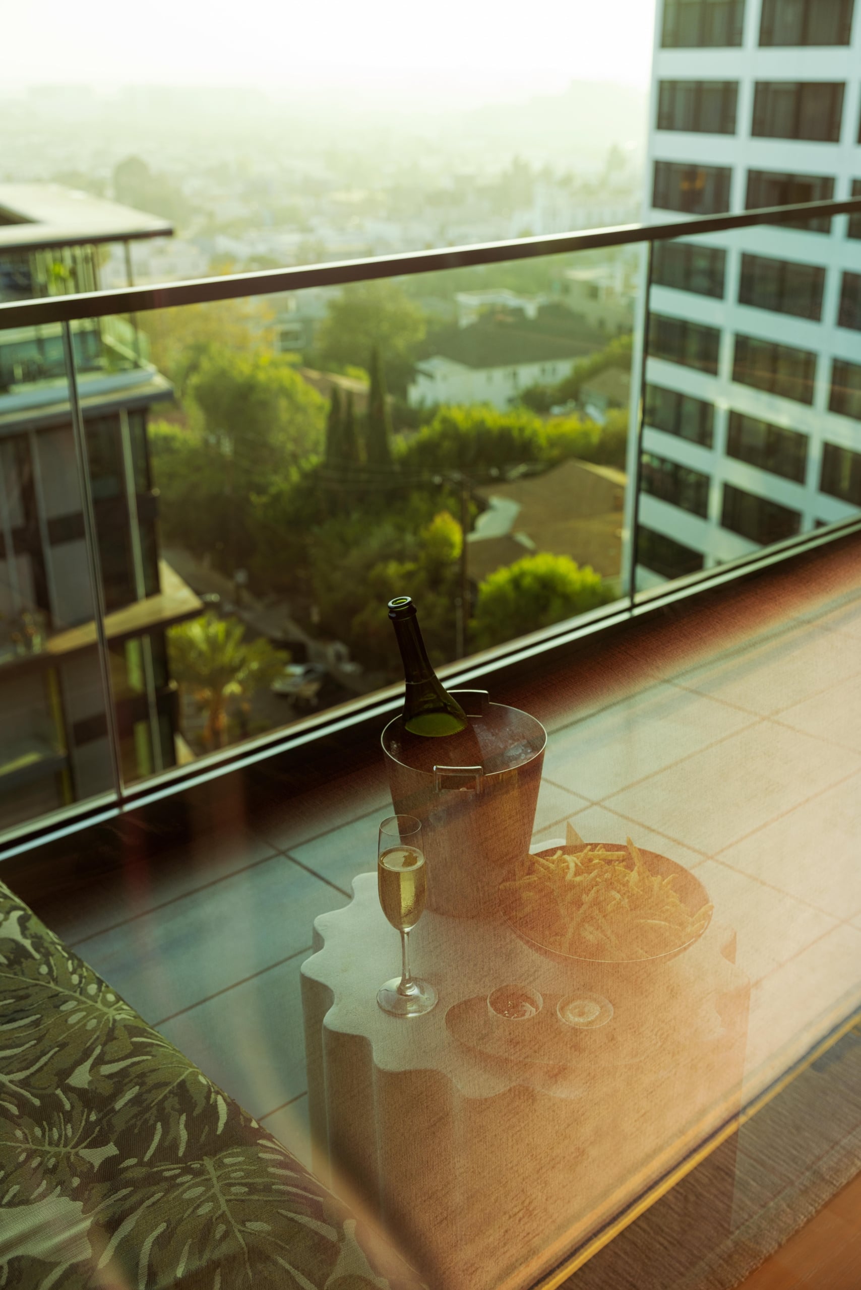 A bottle of champagne on ice and a bowl of fries on a balcony table overlooking a hazy city view.