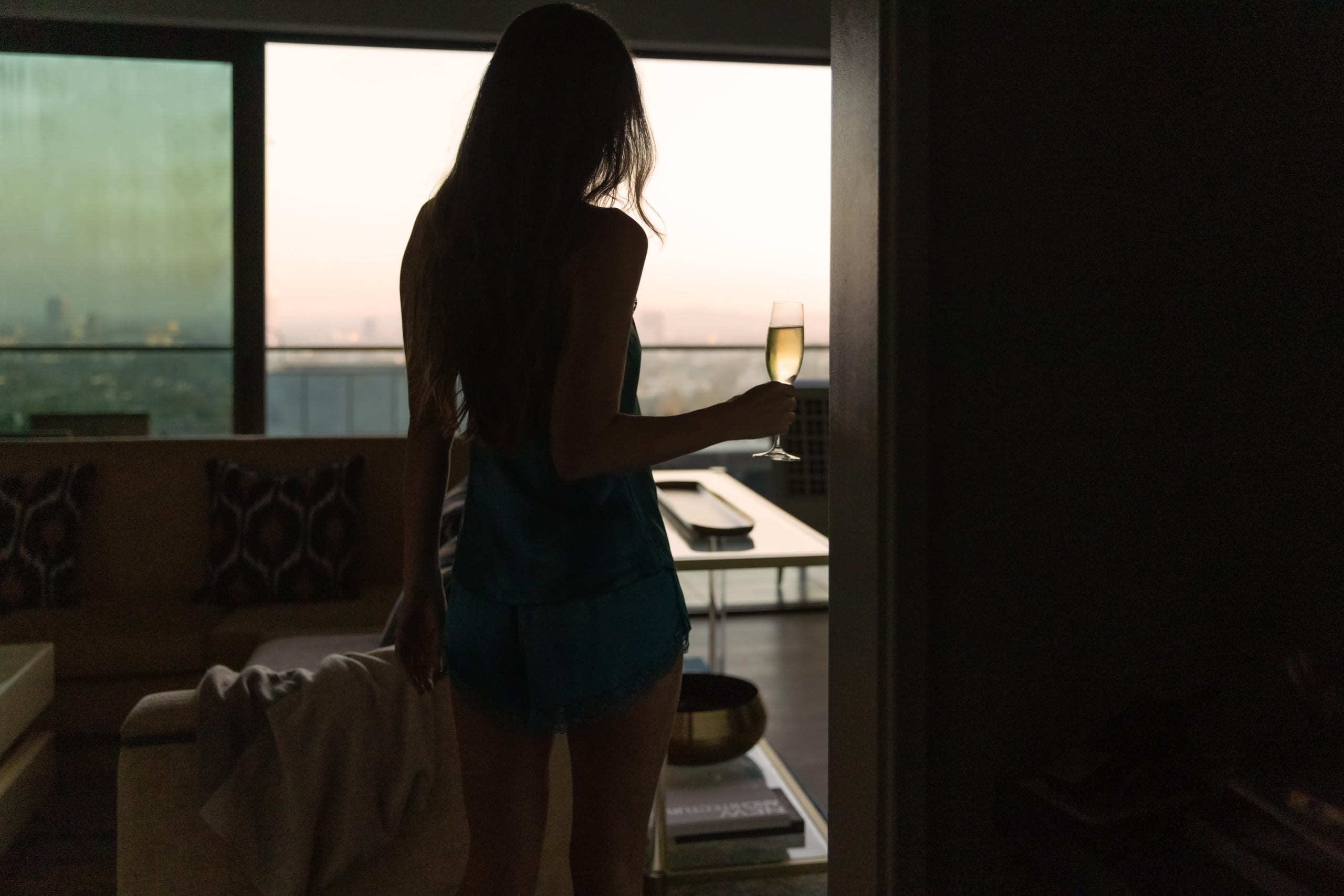 Silhouette of a woman holding a champagne glass in a dark, elegant hotel suite overlooking a bright morning sky.