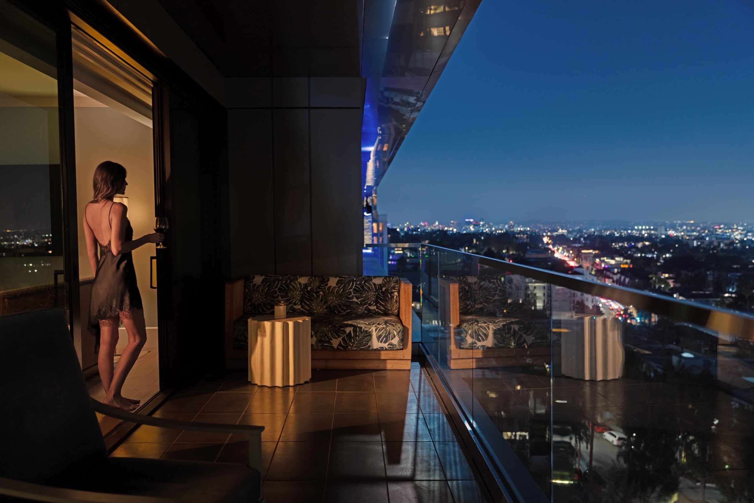 A woman in a black slip dress stands on a hotel balcony at night, looking out over the sparkling city lights.