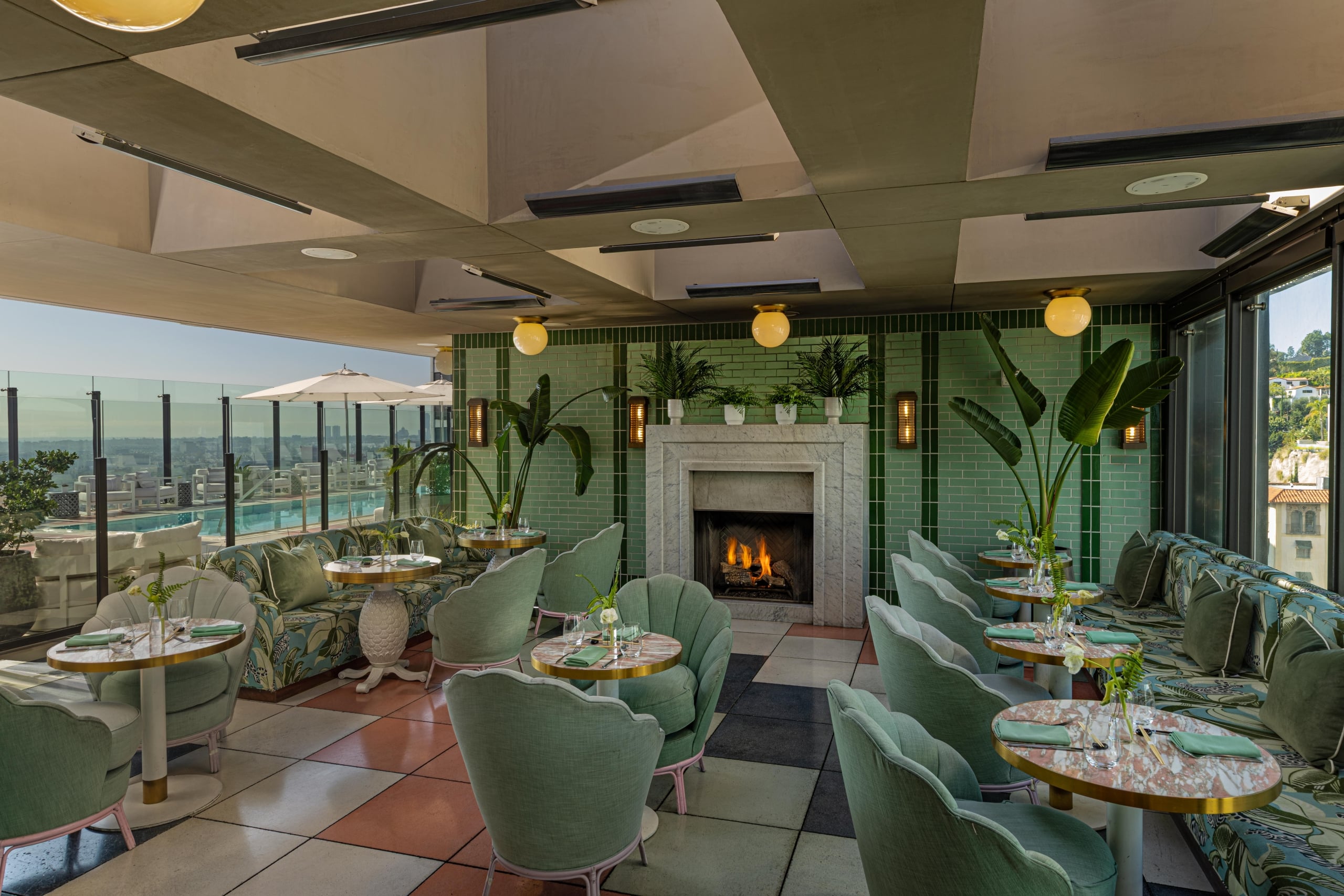 An elegant indoor-outdoor dining room with mint green seating, a marble fireplace, and views of a rooftop pool.