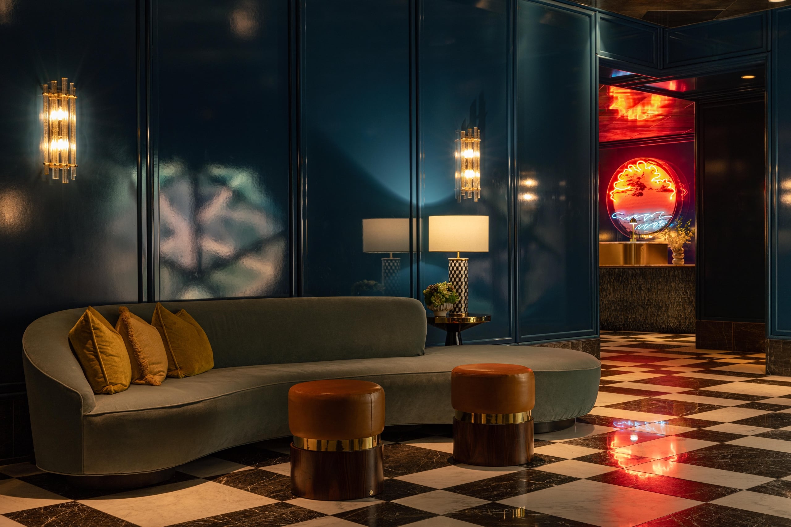 A stylish lounge corner with a curved green sofa, leather ottomans, and a neon sunset sign visible in the background.