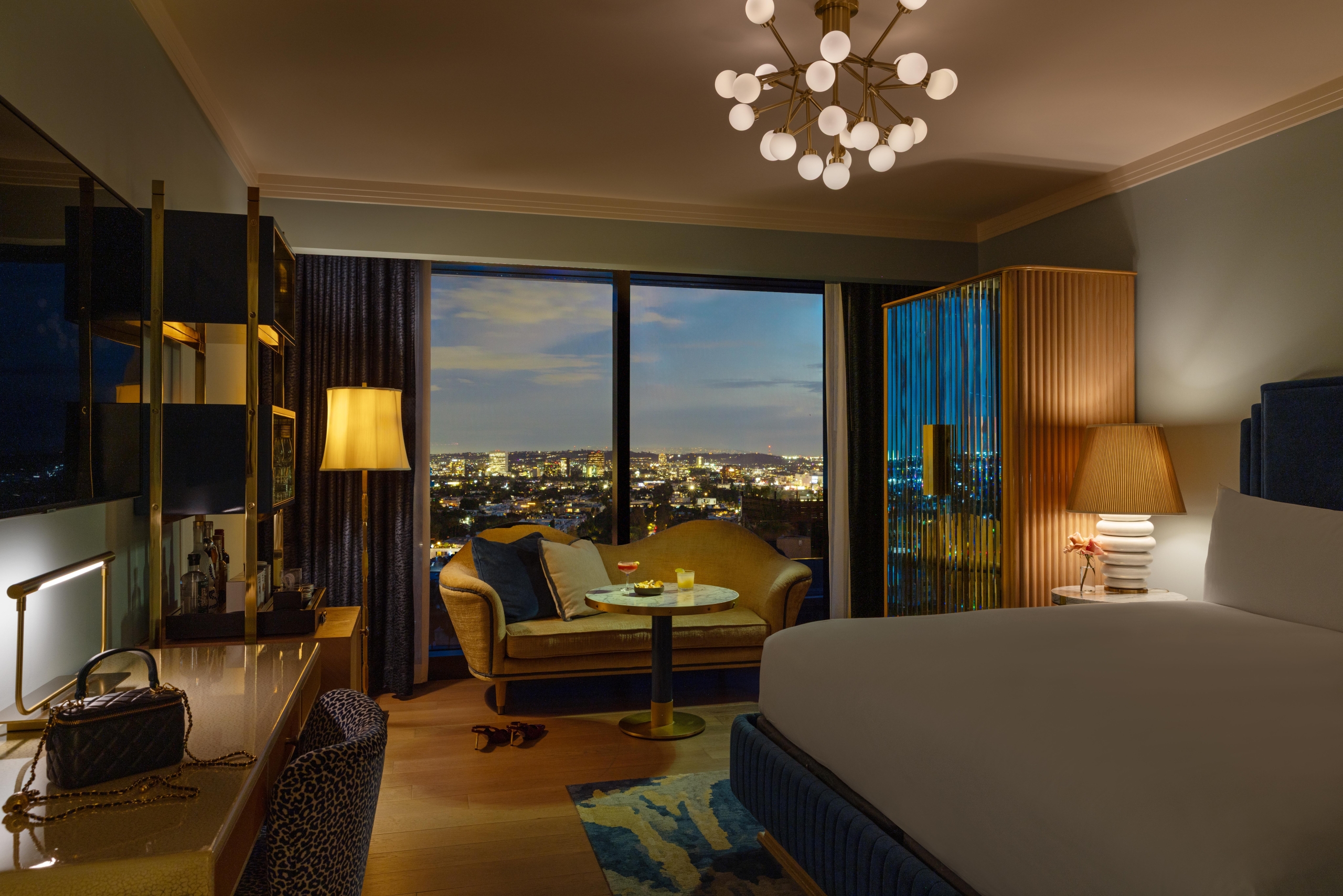 A high-end hotel room at dusk with mid-century modern furniture, a glowing chandelier, and floor-to-ceiling city views.