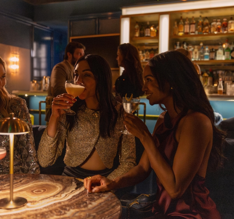 Close-up of three friends enjoying craft cocktails at a dimly lit, upscale bar with warm, ambient lighting.