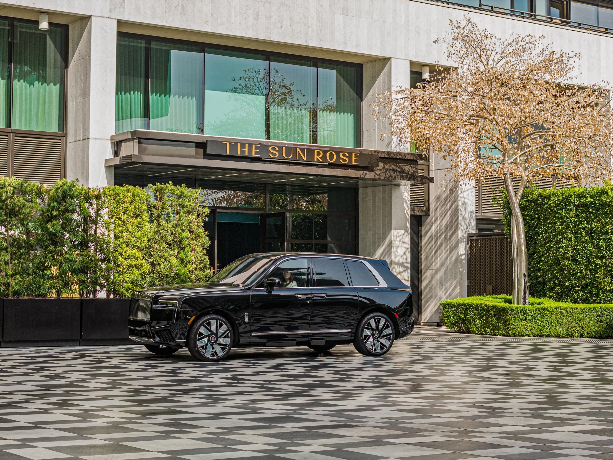 A luxury black SUV parked on the checkered driveway at the elegant entrance of The Sun Rose hotel during the day.