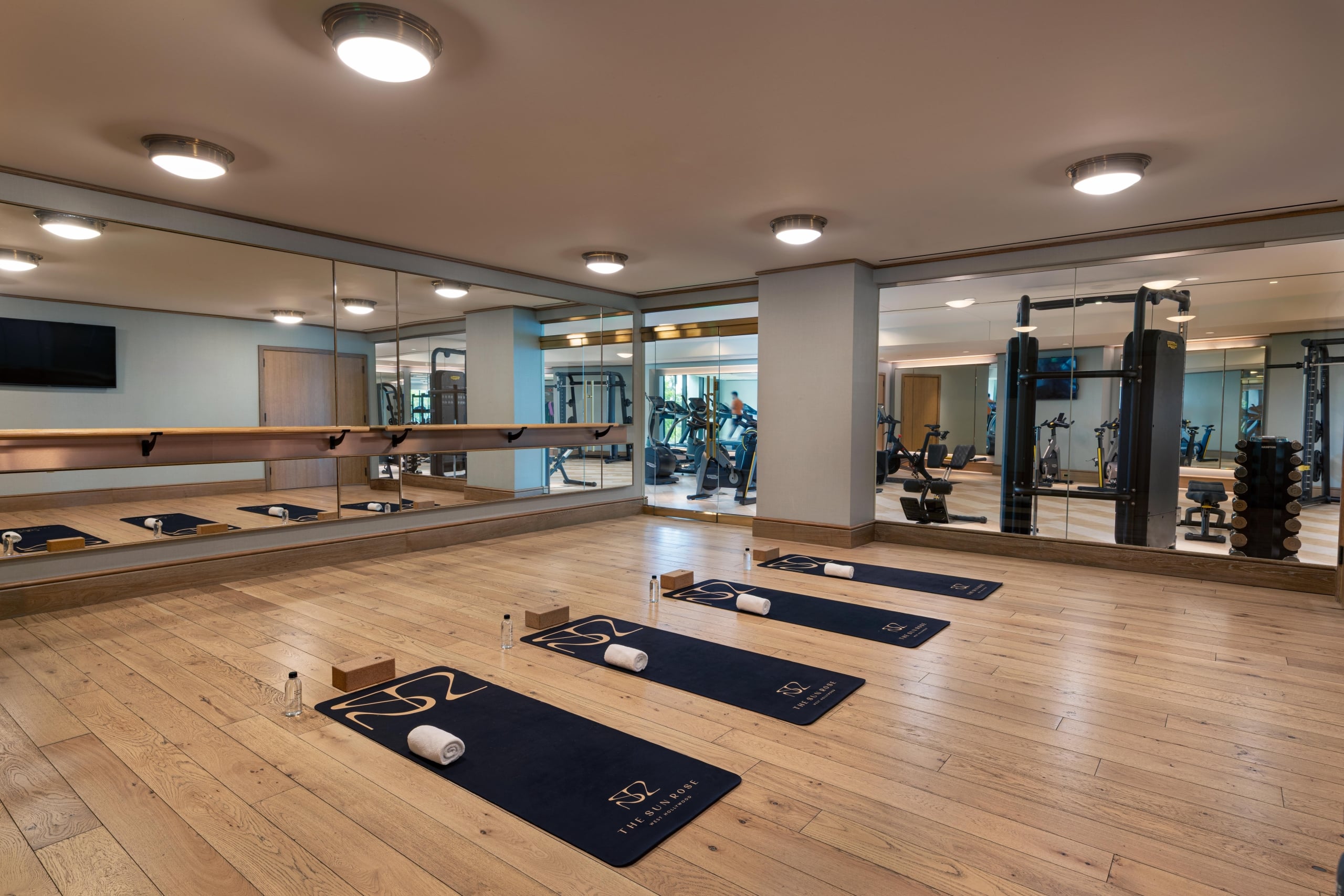 A serene yoga and pilates studio with light wood floors, full-length mirrors, and branded Sun Rose mats ready for use.