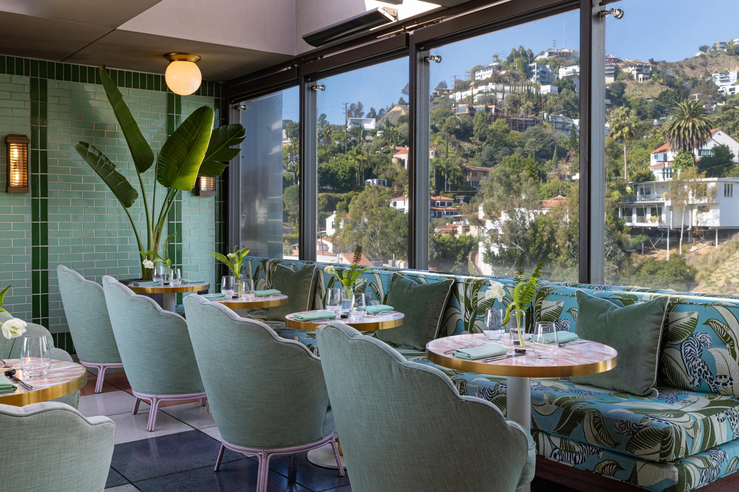An airy rooftop dining area with mint green chairs and floral banquettes overlooking a scenic hillside neighborhood.