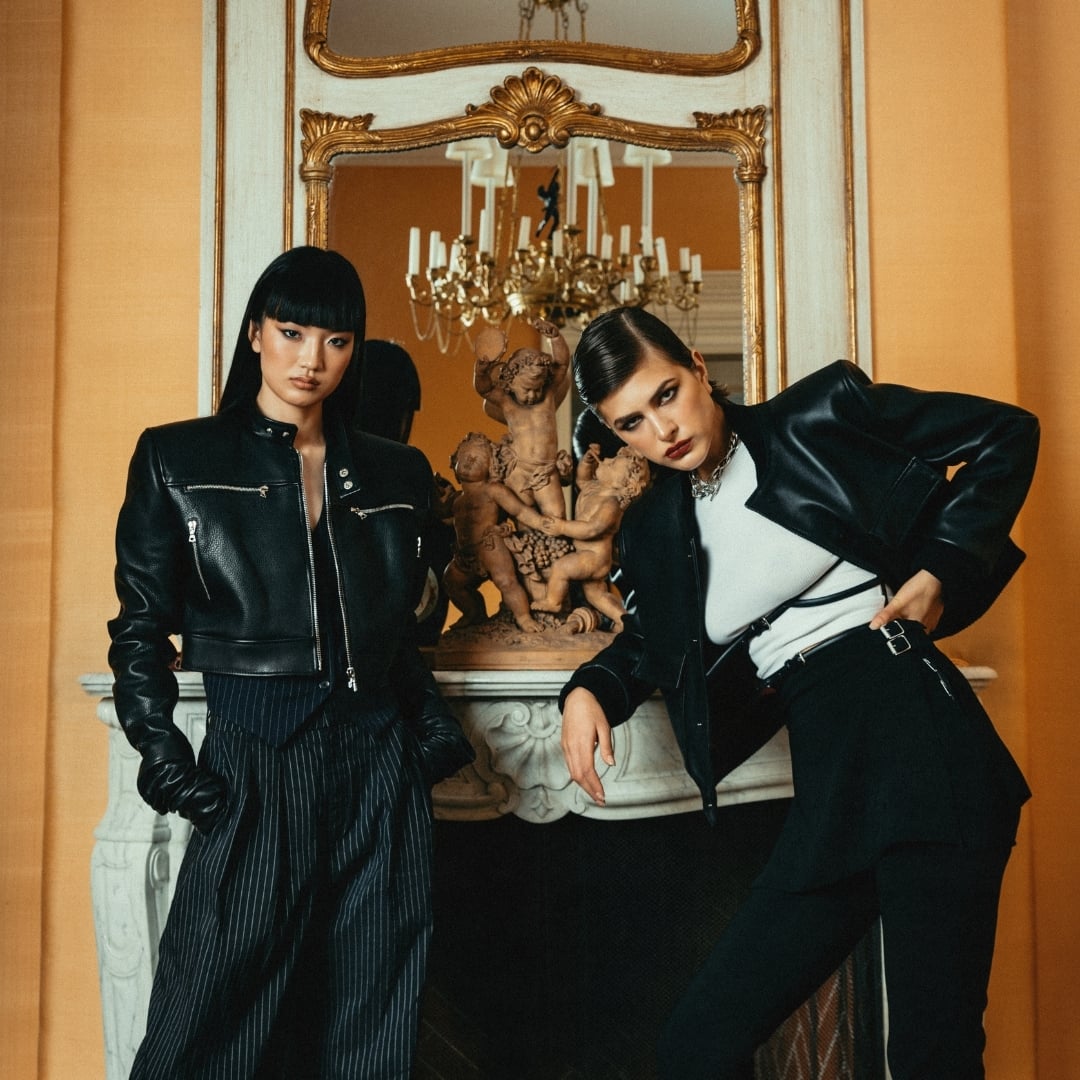Two women wearing customizable leather jackets posing in front of an opulent fireplace with a chandelier reflected in the mirror behind them.