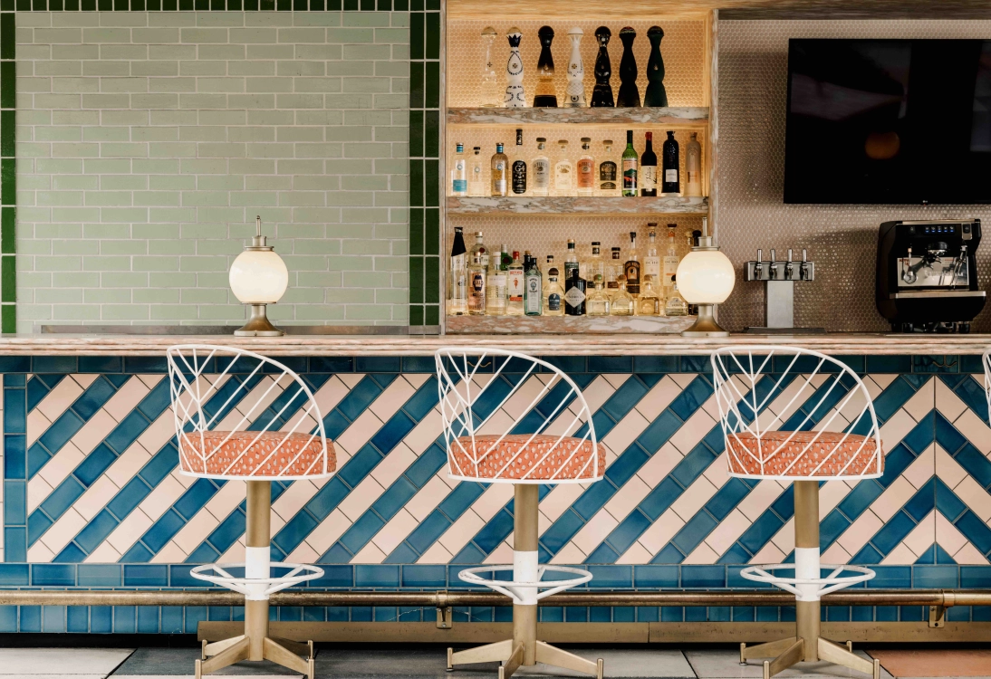 Close up shot of Bar Merois, showing three patterned bar chairs, colorful tiled walls, artwork, an assortment of liquor, and garnishes for drinks.